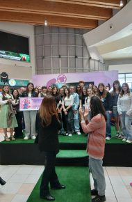 Un gruppo numeroso di studentesse in posa su una pedana con prato sintetico in un centro commerciale; una ragazza tiene il cartellone dell’Eco Fashion Contest, due donne in primo piano applaudono.