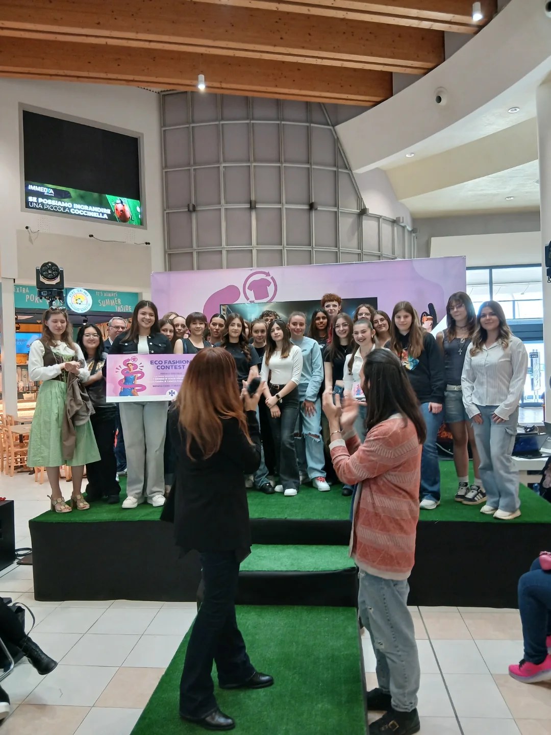 Un gruppo numeroso di studentesse in posa su una pedana con prato sintetico in un centro commerciale; una ragazza tiene il cartellone dell’Eco Fashion Contest, due donne in primo piano applaudono.