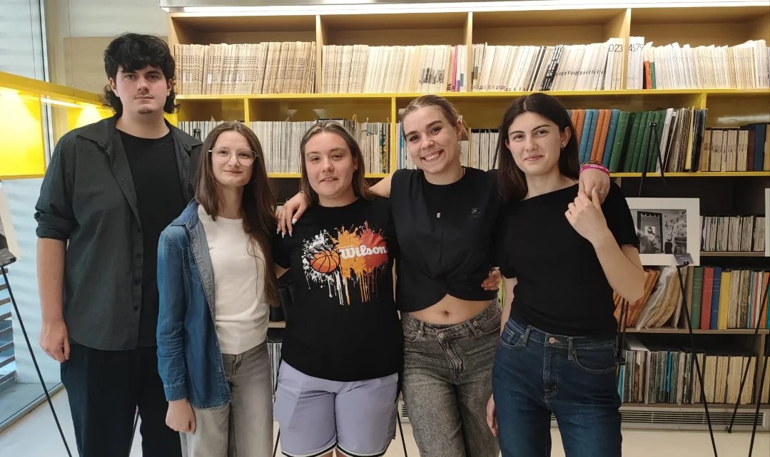 Cinque studentesse e studenti sorridenti in posa davanti agli scaffali di libri della mediateca, accanto a una delle fotografie esposte.