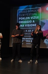 Due studenti e due adulti sul palco di una sala buia; una studentessa parla al microfono davanti allo schermo con il manifesto del Cross-Border Film Festival «Omaggio a una visione».