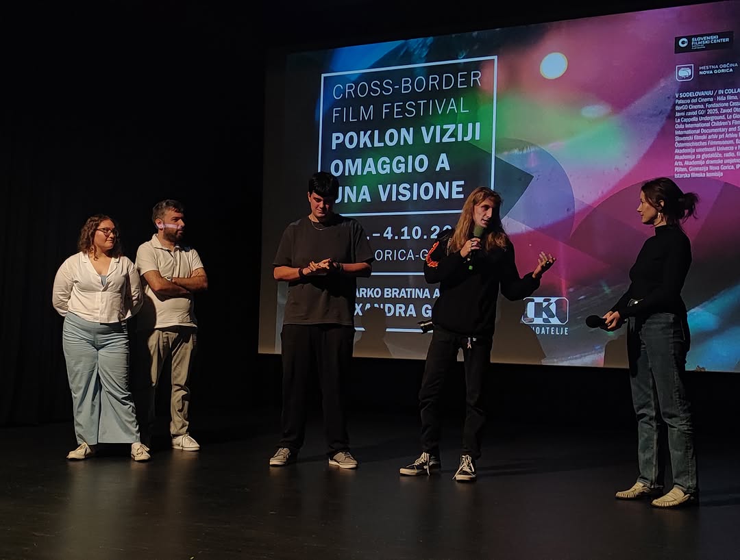 Due studenti e due adulti sul palco di una sala buia; una studentessa parla al microfono davanti allo schermo con il manifesto del Cross-Border Film Festival «Omaggio a una visione».