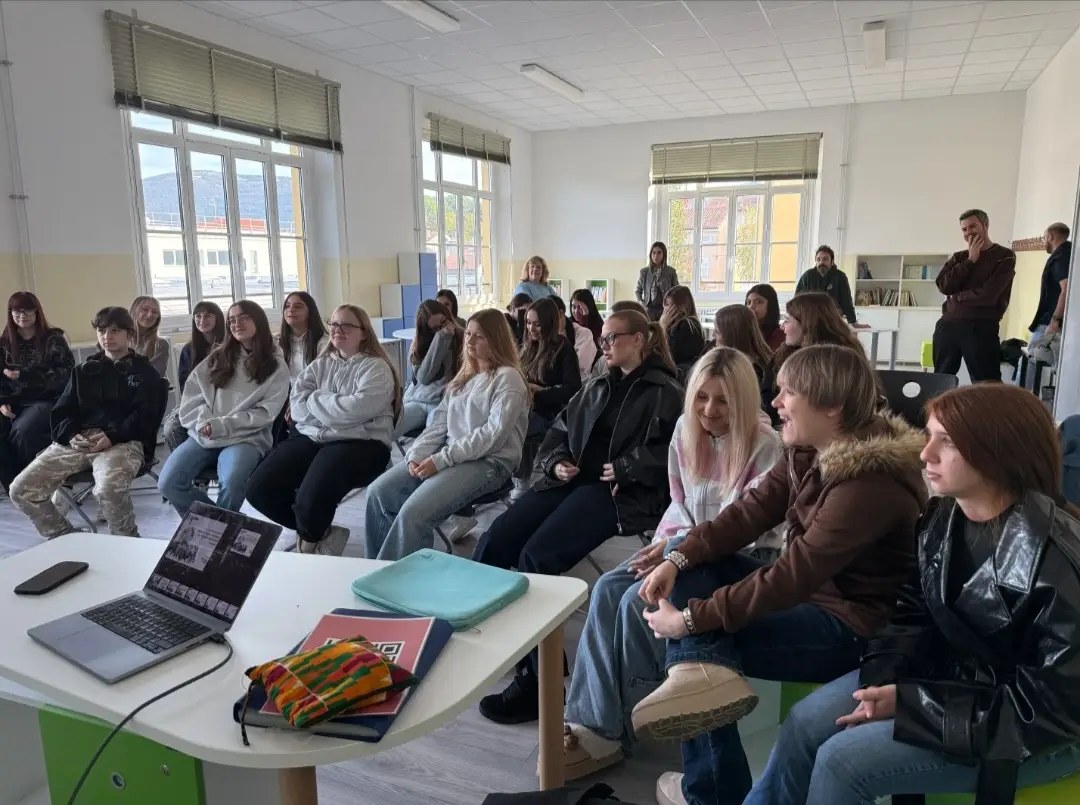 Studentesse e studenti seduti in un’aula luminosa seguono il laboratorio; in primo piano un tavolo con un computer portatile aperto, alcuni docenti in piedi sul fondo.