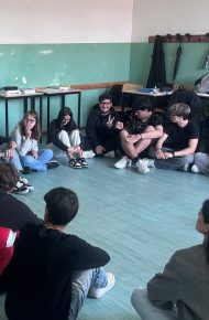 Studentesse e studenti seduti in cerchio sul pavimento di un’aula ascoltano un formatore in piedi.
