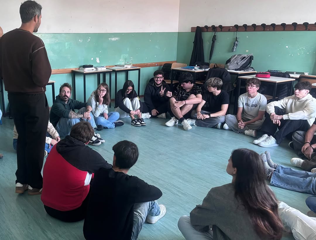Studentesse e studenti seduti in cerchio sul pavimento di un’aula ascoltano un formatore in piedi.